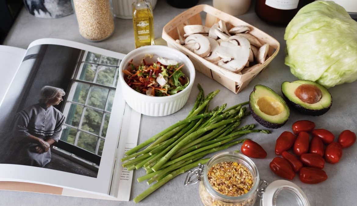 A selection of fresh vegetables and a cookbook in a kitchen setting, promoting healthy cooking.